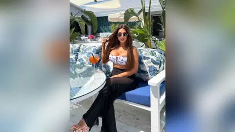 Stylish woman in a white bikini top and black trousers enjoying a cocktail on a sunny tropical patio.