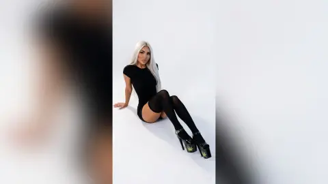 Blonde woman in a black bodysuit and platform heels posing confidently against a white studio background.
