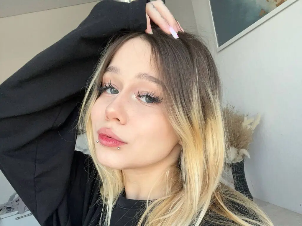 Blonde woman in a black sweatshirt posing for a selfie with a soft, natural aesthetic indoors.