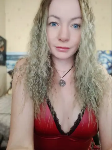 Young woman with long curly blonde hair wearing a red lace camisole and silver necklaces in a cozy bedroom.