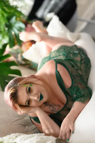 Smiling woman in green lace lingerie relaxing on a white bed in a bright, dreamy indoor setting.