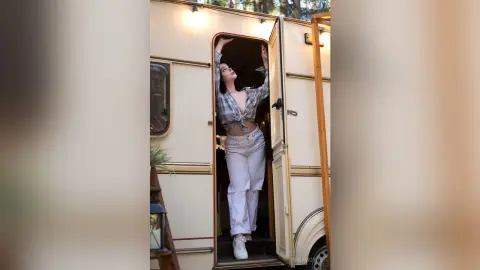 Young woman in a plaid shirt and white trousers posing in a doorway of a vintage camper during a sunny camping trip.