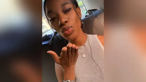 Smiling woman in a grey ribbed tank top blowing a kiss while sitting in a car interior.