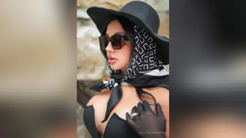 Stylish woman in black sunglasses, sun hat, and patterned headscarf posing outdoors in a chic summer outfit.