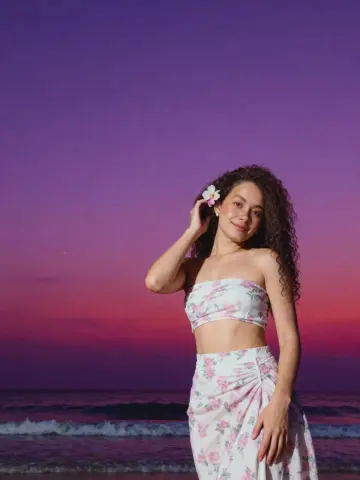 Smiling woman in a floral strapless crop top and skirt posing on a beach during a purple sunset.