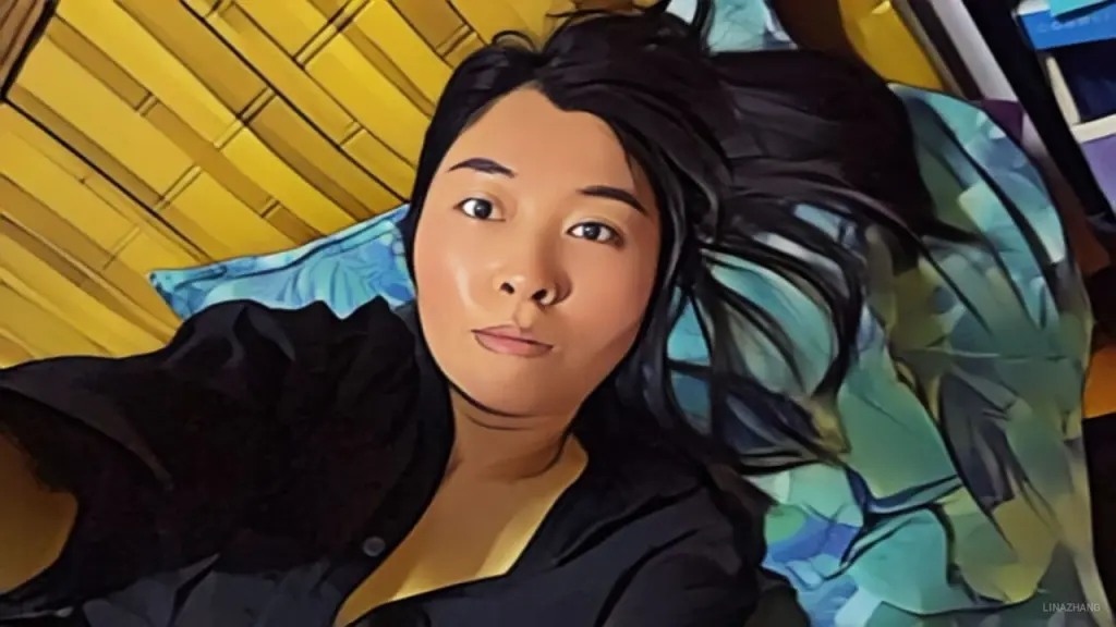 Young woman in a black shirt and glasses sitting on a bed in a bright bedroom setting.