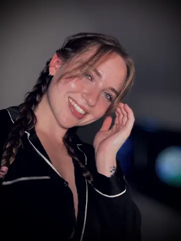 Smiling young woman with braided hair wearing black satin pajamas in a moody, dimly lit indoor setting.