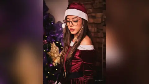 JessieLiu wears a red velvet off-the-shoulder Christmas outfit and a Santa hat while standing near a purple decorated tree.
