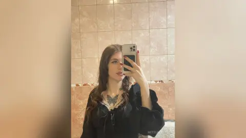 MitsueEastes poses for a mirror selfie wearing a black long-sleeve top with a butterfly tattoo visible on her chest, set against a tiled wall.