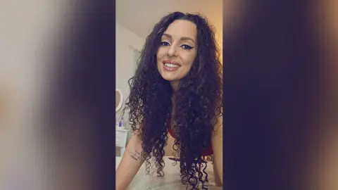 Smiling woman with long curly dark hair in a cozy indoor setting.