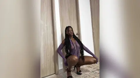 Young Black woman in a sheer purple bodysuit posing confidently in a modern minimalist hallway.