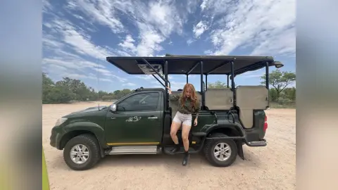 Young woman in casual attire standing by a green safari tour vehicle under a bright cloudy sky.