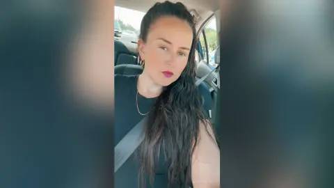 Young woman with long dark hair wearing a black t-shirt and gold necklace sitting in a car.