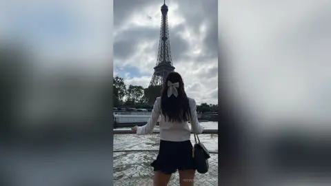 Woman in white blouse and black skirt standing by the Seine river facing the Eiffel Tower in Paris.