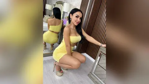 Glamorous woman in a tight yellow mini dress posing in a modern bedroom setting.