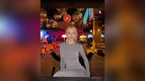 Elegant blonde woman in a grey long-sleeve dress sitting in a moody, warm-lit luxury lounge with glowing orb lights.