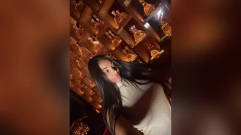 Young woman with long dark hair in a white ribbed top posing in a warm, glowing luxury lounge setting.