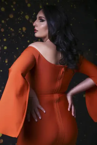 Elegant woman in an orange off-the-shoulder long sleeve dress posing against a dark sparkly background.