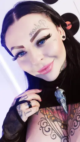 Smiling woman with forehead tattoos and black hair wearing a gothic skull necklace in a bright room.