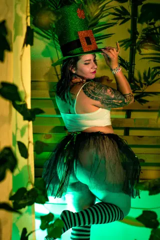 Woman in leprechaun costume with green top hat and black tutu posing in a lush garden setting.