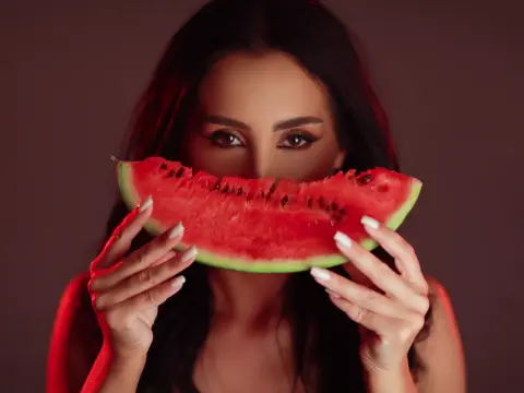 Woman with dark hair holding a fresh watermelon slice in front of her face against a moody red background.