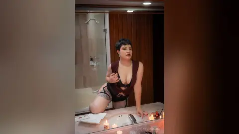 Curvy woman in a black lace bodysuit and grey shorts posing in a modern bathroom with warm lighting.
