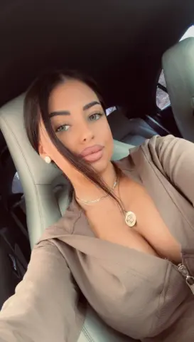 Young woman with long dark hair wearing a beige loungewear outfit and gold necklace sitting in a car.