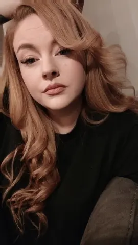 Young woman with wavy strawberry blonde hair wearing a black t-shirt in a soft, moody indoor setting.