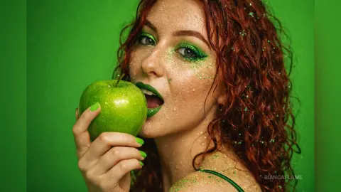 Redheaded woman with green glitter makeup biting a green apple against a vibrant green background.