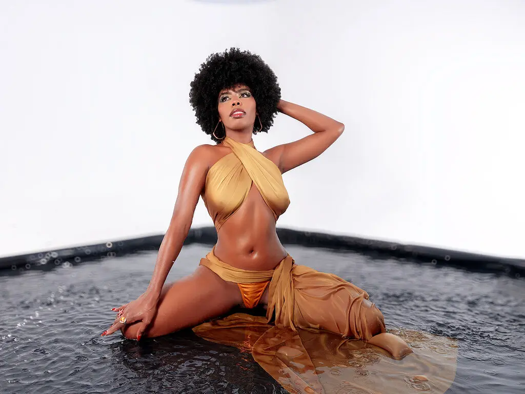 Elegant woman in a gold halter swimsuit sitting on a dark water surface against a white background.