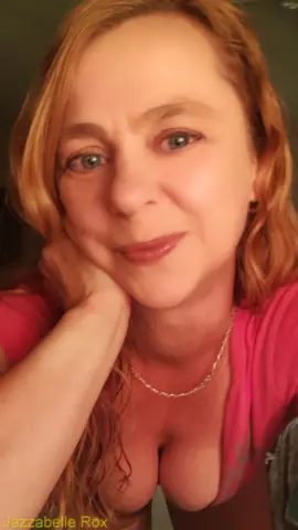 Smiling blonde woman in a pink top wearing a gold necklace in a warm indoor setting.