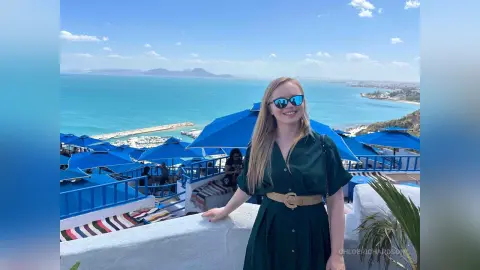 Smiling blonde woman in a green dress and sunglasses enjoying a sunny coastal view at a blue seaside resort.