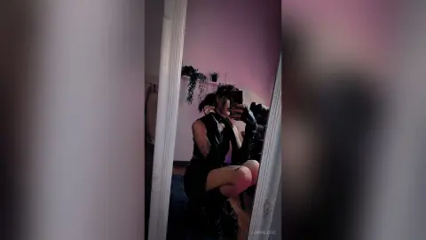 LorenLoiss poses in a black outfit, holding a phone to take a mirror selfie in a room with pink lighting.
