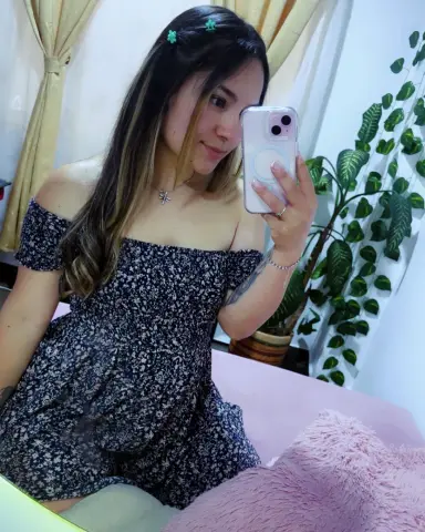 Young woman in a floral off-the-shoulder romper taking a mirror selfie in a bright room with indoor plants.