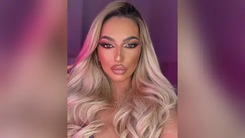 Blonde woman with glamorous winged eyeliner and wavy hair posing in soft pink studio lighting.