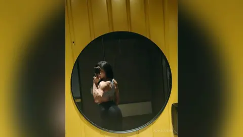 Fit woman in grey sports bra taking a mirror selfie in a modern yellow gym setting.