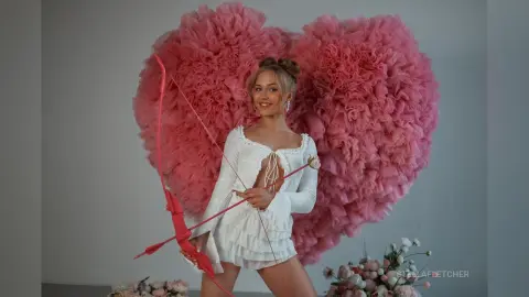 StellaFletcher poses in a white long-sleeve top against a large pink heart-shaped ruffly backdrop while holding a red bow.