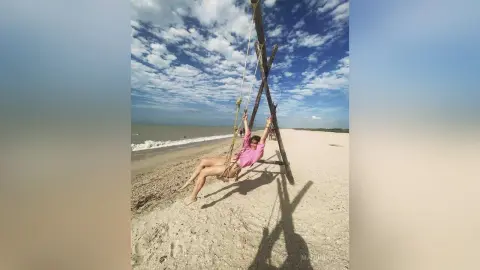 Happy woman in a pink shirt swinging on a wooden swing at a sunny tropical beach under a blue sky.