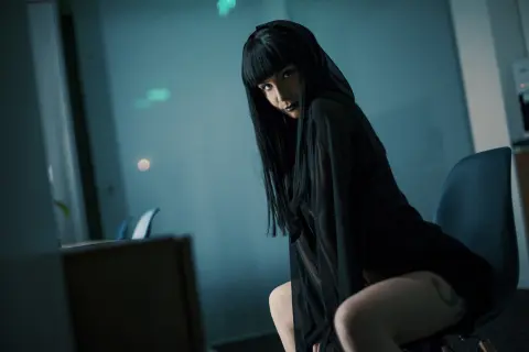 Mysterious woman in a sheer black outfit sitting in a moody, dimly lit modern office setting.