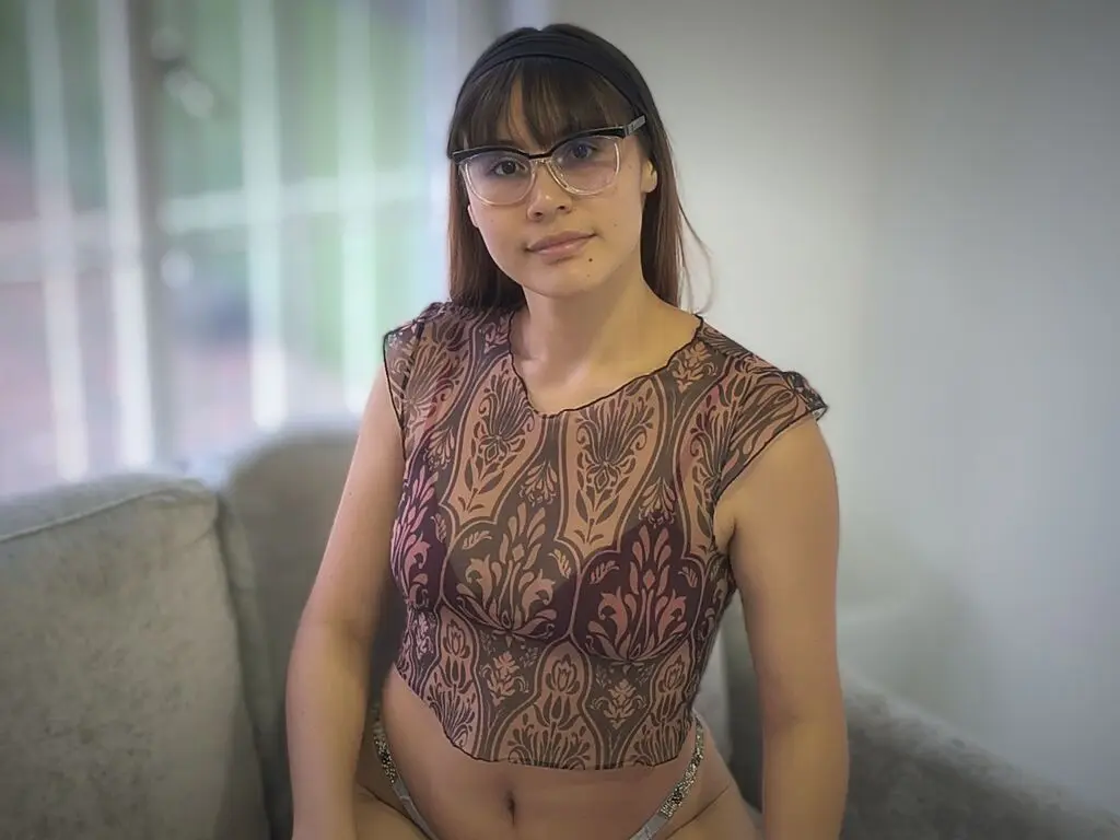 Young woman with glasses in a patterned brown sheer top sitting on a sofa in a bright room.