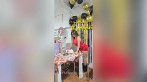 Young woman in a red mini dress celebrating a birthday with cake and gold balloons in a festive indoor setting.