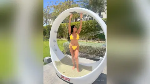 MissyJolie stands inside a white circular swing wearing a yellow bikini, raising her arms above her head.