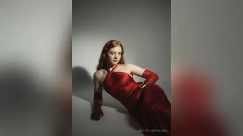 Elegant redhead woman in a glamorous red satin gown and matching gloves posing against a minimalist gray backdrop.
