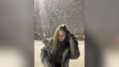 Young woman in a dark puffer jacket standing in a heavy snowfall at night with a moody atmosphere.