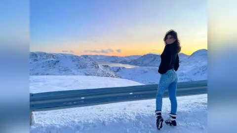 AnaMathers stands outdoors in a snowy mountain landscape wearing blue jeans and black top.
