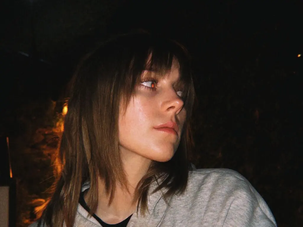 Young woman with brown hair in a grey sweatshirt looking away in moody, low-light nighttime setting.
