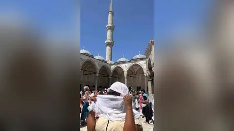 Tourist in a white headscarf exploring a historic mosque courtyard under a bright blue sky.