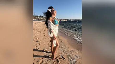 MissyJolie stands on a sandy beach wearing a light blue bikini top and a white mesh cover-up with a white flower in her hair.