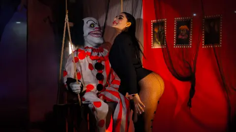 Woman in black blazer and fishnets posing with a creepy clown prop in a spooky, red-lit haunted house setting.