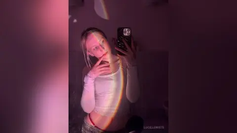 Young woman in a white tank top taking a moody selfie in soft pink lighting.
